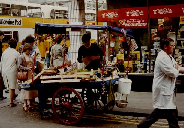 A view of a Street Trader on Brunswick Place Newcastle upon Tyne taken in 1983. The photograph shows a Street Trader serving customers from his fruit barrow. F.W. Woolworth's store can be seen behind the barrow to the right and buses travelling down Northumberland Street to the left.