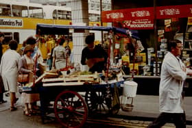 A view of a Street Trader on Brunswick Place Newcastle upon Tyne taken in 1983. The photograph shows a Street Trader serving customers from his fruit barrow. F.W. Woolworth's store can be seen behind the barrow to the right and buses travelling down Northumberland Street to the left.